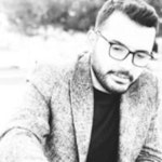 Bu, siyah çerçeveli gözlük ve lacivert blazer ceket giyen bir adamın fotoğrafı. Kısa, koyu kahverengi saçları ve bakımlı bir keçi sakalı var. Yüz ifadesi nötr ve doğrudan kameraya bakıyor. Ceketin altına üst düğmesi açık beyaz bir elbise gömleği giymiştir. Profesyonel bir ortamda, muhtemelen bir şirket ofisinde duruyor gibi görünüyor. Beden dili kendine güven ve sofistike bir hava taşıyor. Profesyonel görünümünü tamamlayan siyah deri bir kemer ve siyah elbise ayakkabıları giyiyor.
