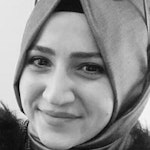 Resimdeki kadın, omuzlarından sarkan ve göğsünü örten beyaz ve mavi desenli bir eşarp takıyor. Kahverengi saçlarını arkaya toplamış ve kendinden emin bir ifadeyle dümdüz karşıya bakıyor. Gözleri kahverengi ve teni açık renk. Beyaz bir üst ve mavi kot pantolon giyiyor. Boynunda tek mavi taşlı bir kolye var. Resmin arka planı açık mavi ve gri karışımı. Kadın, uzakta birkaç ağacın bulunduğu açık bir alanda duruyor.