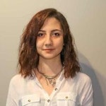 Resimdeki kadının uzun sarı saçları sırtına kadar uzanıyor. Önünde küçük bir logo bulunan beyaz bir tişört giyiyor. Yüzünde kendinden emin bir ifadeyle doğrudan kameraya bakıyor. Gözleri koyu kahverengi ve dudakları hafif bir gülümseme şeklinde kıvrılmış. Cildi pürüzsüz ve kremsi bir tonda ve yanaklarında pembe bir parlaklık var. Basit bir gümüş kolye ve bir çift küçük halka küpe takıyor. Kollarını önünde kavuşturmuş ve ellerini birbirine kenetlemiş. Beyaz duvarları ve mavi mobilyaları olan aydınlık ve havadar bir odada duruyor.