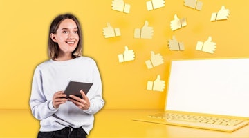 A woman is pictured smiling while holding a tablet computer. She is sitting in front of a laptop with a white screen, and her hands are positioned on the tablet, which has a brightly colored background. She has a bright yellow background to her, and her eyes are focused on the tablet. In the background, there is a group of thumbs-up symbols in different colors, including a white one on the top right and a yellow one on the bottom left. The woman is wearing a suit, and her hair is tied back. Her expression is one of joy and contentment.