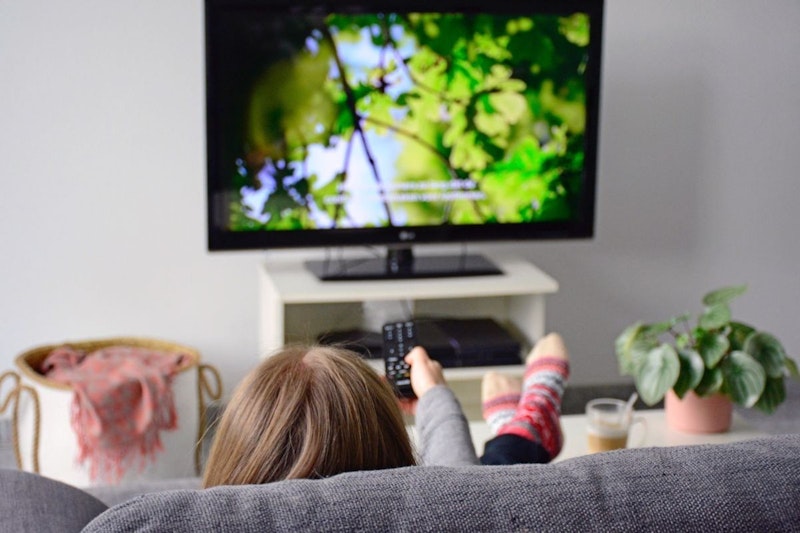 Bir kişi televizyonun önündeki kanepede oturuyor, bacak bacak üstüne atmış ve elleri kucağında. Televizyon yakın çekim ve hafif bulanık, açık mavi renkte. Televizyonun sağında bir masanın üzerinde bir saksı bitkisi var. Ön planda, yine hafifçe bulanıklaştırılmış, pipetli bir bardak var. Arka planda bir kumaş yüzeyinin yakın çekimi görülüyor. Kişinin yüzü odaktadır ve ayaklarının dibinde bir çantanın yakın çekimi görülmektedir. Daha uzakta, bir kişinin kolu bulanık ve bir düğmeye basıyor. Renkli bir çorap giyen ayakları da görülebiliyor. Bu görüntü bir rahatlama ve eğlence anını yakalıyor.
