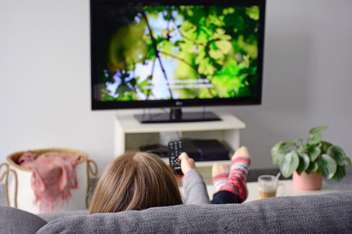 Bir kişi televizyonun önündeki kanepede oturuyor, bacak bacak üstüne atmış ve elleri kucağında. Televizyon yakın çekim ve hafif bulanık, açık mavi renkte. Televizyonun sağında bir masanın üzerinde bir saksı bitkisi var. Ön planda, yine hafifçe bulanıklaştırılmış, pipetli bir bardak var. Arka planda bir kumaş yüzeyinin yakın çekimi görülüyor. Kişinin yüzü odaktadır ve ayaklarının dibinde bir çantanın yakın çekimi görülmektedir. Daha uzakta, bir kişinin kolu bulanık ve bir düğmeye basıyor. Renkli bir çorap giyen ayakları da görülebiliyor. Bu görüntü bir rahatlama ve eğlence anını yakalıyor.