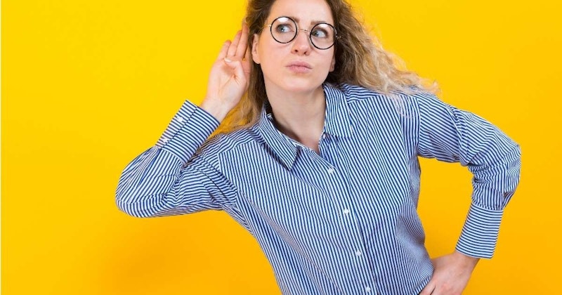 Genç bir kadın elini kulağına götürmüş, gözleri uzaktaki bir şeye odaklanmış. Dudakları hafifçe ayrılmış ve ifadesi ciddi. Uzun, koyu renk saçları yüzünden geriye doğru toplanmış. Teni soluk ve giysileri siyah. Duruşu kendinden emin ve beden dili tetikte olduğunu gösteriyor. Sanki bir şey dinliyormuş gibi derin düşüncelere dalmış gibi görünüyor. Arka plan odak dışı ve sadece küçük ayrıntılar görülebiliyor.