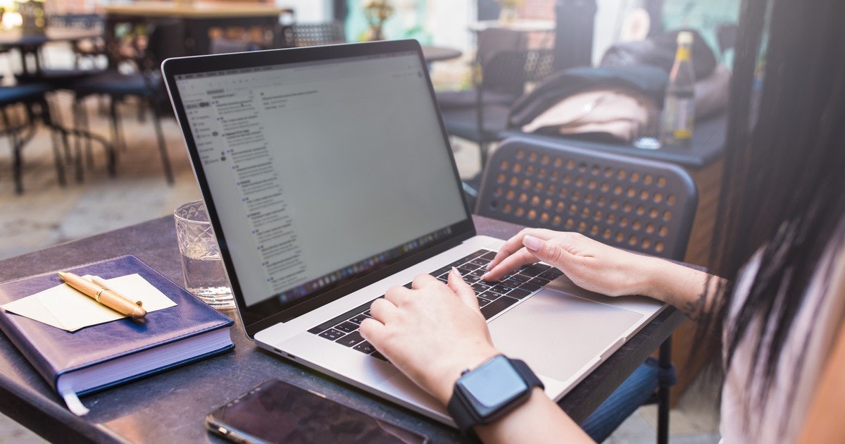 Bir kişi dizüstü bilgisayarın önünde oturmuş, klavyede yazıyor. Bir elini trackpad'in üzerine koymuş, diğer eliyle de yazmaya devam ediyor. Dizüstü bilgisayar açık ve ekran aydınlatılmış, çeşitli sekmeler ve pencereler gösteriliyor. Kişinin parmağında bir saat ve bir yüzük var ve yakınındaki bir kitabın üzerinde bir kalem duruyor. Arka plan hafifçe bulanıktır ve ön planda odakta olan bir bardak vardır. Kişi görevine odaklanmıştır ve dizüstü bilgisayar onun tek yoldaşıdır.