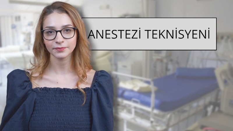 Gözlük ve mavi gömlek giyen bir kadın. Kızıl saçları var ve yüzü yakından görünüyor. Boynu da yakın plandadır ve geniş bir gülümsemesi vardır. Gözleri görülebiliyor ve gözlük camları ışığı yansıtıyor. Mavi gömlek düz bir renk ve dar. Gömleğin alt kısmı kot pantolonunun içine sokulmuştur. Arka planda siyah metinli beyaz bir tabela var. Arka planda bir hastane yatağının bulanık bir görüntüsü de var.