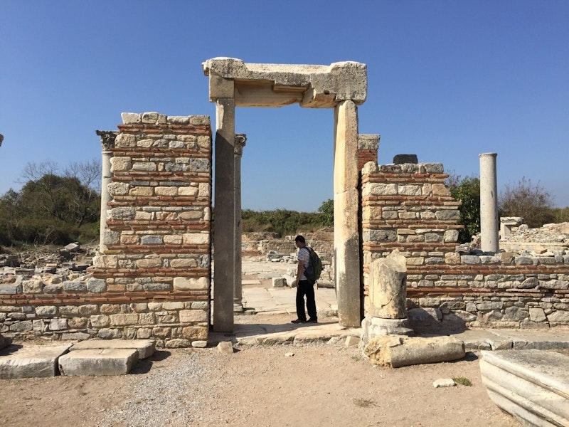 Bir adam büyük bir taş kemerin önünde duruyor. Sırt çantası takıyor ve yüzünde sakin, rahat bir ifade var. Arkasında üzerinde siyah bir leke olan beyaz bir sütun var. Sağında taş sütunlu bir taş duvar ve arka planda bir tarlada bir grup ağaç var. Gökyüzü mavidir ve güneş parlamaktadır. Taş kemer birkaç büyük taş levhadan oluşuyor ve karmaşık bir tasarıma sahip. Adam kemerin ortasında durmakta ve dramatik bir görüntü oluşturmaktadır.