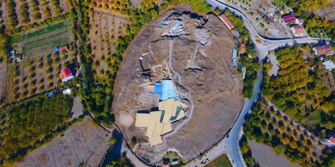 Bu, bir yol ve ağaçların bulunduğu geniş bir alanın havadan görünümüdür. Yol dolambaçlı ve her iki tarafı da uzun, yeşil ağaçlarla kaplı. Yolun ilerisinde, ağaçlar ve yeşilliklerle çevrili büyük bir bina görülüyor. Sol tarafta, tarlaları ve bir eviyle bir çiftlik görülebilir. Daha arka planda bir orman, ön planda ise kırmızı bir kutunun bulanık görüntüsü görülüyor. Genel olarak, geniş alan ağaçlar ve bitkilerle dolu, manzara boyunca dağılmış birkaç bina ve yol var.