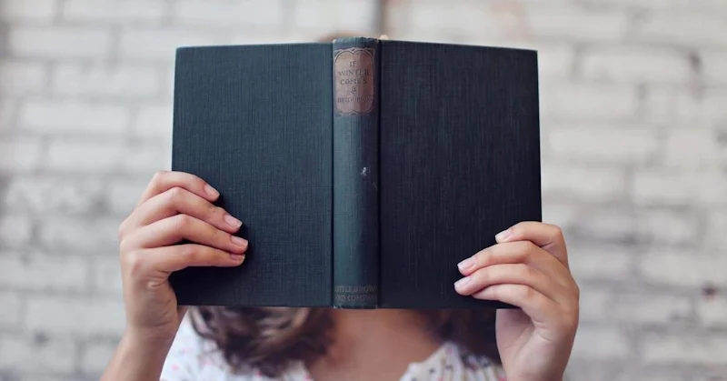 Bir kişi elinde bir kitapla yüzünü gizlerken görülüyor. Arkasında duvar olan bir odada ayakta durmakta, kolları ve kitap önünde durmaktadır. Kişi beyaz bir gömlek ve koyu renkli bir pantolon giymektedir. Kitap açık renkli bir kapağa sahiptir ve kişinin elinde işaret ve orta parmaklarıyla tutulmaktadır. Kitap kişinin yüzüne doğru tutulmuş ve neredeyse tamamen kapatılmıştır. Kişinin yüz ifadesi görünmüyor. Odanın arka planı çok minimal, sadece küçük bir masa ve bir sandalye görünüyor.