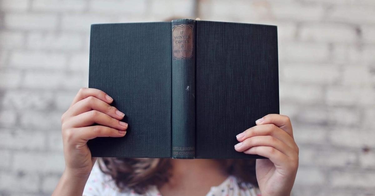 A person is seen holding a book, obscuring their face with it. They are standing in a room with a wall behind them, and their arms and the book in front of them. The person is wearing a white shirt and dark colored pants. The book has a light colored cover and is held in the person's hands with their index and middle fingers. The book is held up to the person's face, covering it almost completely. The person's expression is not visible. The background of the room is very minimal, with only a small table and a chair visible.