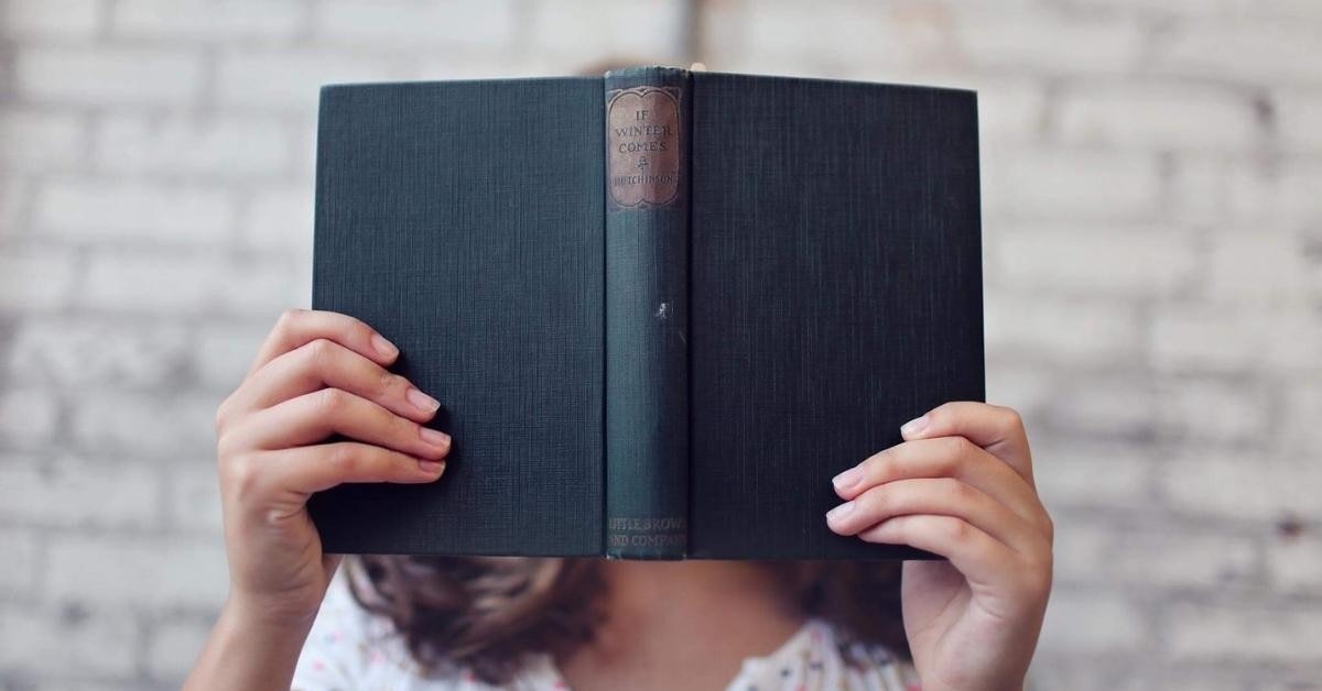 A person is seen holding a book, obscuring their face with it. They are standing in a room with a wall behind them, and their arms and the book in front of them. The person is wearing a white shirt and dark colored pants. The book has a light colored cover and is held in the person's hands with their index and middle fingers. The book is held up to the person's face, covering it almost completely. The person's expression is not visible. The background of the room is very minimal, with only a small table and a chair visible.