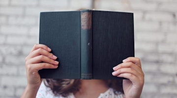 A person is seen holding a book, obscuring their face with it. They are standing in a room with a wall behind them, and their arms and the book in front of them. The person is wearing a white shirt and dark colored pants. The book has a light colored cover and is held in the person's hands with their index and middle fingers. The book is held up to the person's face, covering it almost completely. The person's expression is not visible. The background of the room is very minimal, with only a small table and a chair visible.