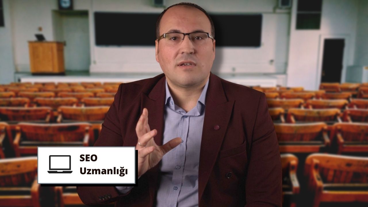 Şık, koyu renk bir takım elbise giymiş bir adam bir konferans salonunun ortasında durmaktadır. Gözlük takmış ve ciddi bir ifadeyle dosdoğru önüne bakıyor. Elleri iki yanındadır. Görüntünün odağında o var ve arka plan hafifçe bulanık. Bir iş adamı gibi görünüyor ve arkasındaki siyah yazılı dikdörtgen beyaz tabela bunu gösteriyor olabilir. Sağ üst köşede bir sandalye ve sol alt köşede bir masa var, her ikisi de odak dışı.