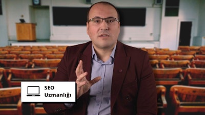 Şık, koyu renk bir takım elbise giymiş bir adam bir konferans salonunun ortasında durmaktadır. Gözlük takmış ve ciddi bir ifadeyle dosdoğru önüne bakıyor. Elleri iki yanındadır. Görüntünün odağında o var ve arka plan hafifçe bulanık. Bir iş adamı gibi görünüyor ve arkasındaki siyah yazılı dikdörtgen beyaz tabela bunu gösteriyor olabilir. Sağ üst köşede bir sandalye ve sol alt köşede bir masa var, her ikisi de odak dışı.