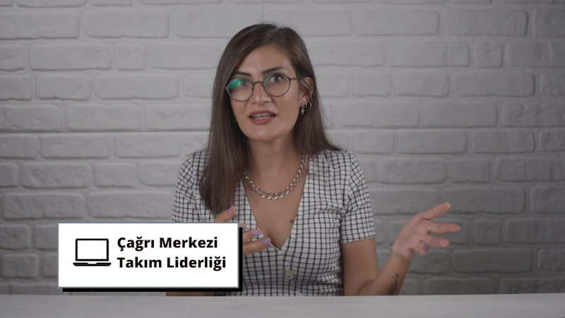 Gözlüklü bir kadın bir duvarın önünde duruyor, gülümsüyor ve elinde siyah yazılı beyaz bir tabela tutuyor. Kareli bir gömlek giymiş ve boynunda bir kolye var. Gözleri gözlüklerin arkasından görülebiliyor ve kahverengi saçları düşük bir at kuyruğu şeklinde toplanmış. Sağ eliyle tabelayı kameraya doğru tutuyor ve yüzünde kararlı bir ifade var. İfadesi kendinden emin ve duruşu güçlü.