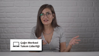 Gözlüklü bir kadın bir duvarın önünde duruyor, gülümsüyor ve elinde siyah yazılı beyaz bir tabela tutuyor. Kareli bir gömlek giymiş ve boynunda bir kolye var. Gözleri gözlüklerin arkasından görülebiliyor ve kahverengi saçları düşük bir at kuyruğu şeklinde toplanmış. Sağ eliyle tabelayı kameraya doğru tutuyor ve yüzünde kararlı bir ifade var. İfadesi kendinden emin ve duruşu güçlü.