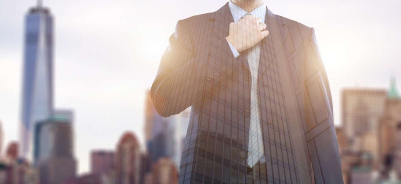 Lacivert takım elbise giyen bir adam, parlak beyaz arka plana sahip bir duvarın karşısında durmaktadır. Sol elinde kravatının düğümünü tutuyor, sağ elini ise cebine sokmuş. Yüzü odaklanmış ve kararlı, ancak gözleri aşağıya bakıyor. Takım elbisesi, geniş omuzlarını ve ince belini vurgulayacak şekilde dikilmiş ve tam oturmuştur. Takım elbisesinin altına, üst iki düğmesi açık beyaz bir gömlek giymiştir. Açık kahverengi saçları geriye taranmıştır ve çene hattı keskin ve köşelidir. Duruşu kendinden emin ve iddialı ve çerçevede uzun duruyor. Kravatı koyu mavi renkte ve ince bir desene sahip ve mükemmel bir şekilde düğümlenmiş.