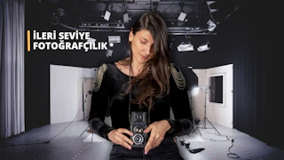 Uzun mor elbiseli bir kadın beyaz bir duvarın önünde durmuş, elinde siyah ve gümüş renkli bir fotoğraf makinesi tutmaktadır. Yüzü kameranın flaşıyla aydınlanmış, koyu renk saçları yüzünden geriye doğru toplanmıştır. Yüz ifadesi ciddi ve odaklanmış bir şekilde kameraya bakmaktadır. Arka planda, yerde beyaz kare bir nesne ve siyah bir kordon görülüyor. Sahne, kadının arkasındaki bir perdeyle çerçevelenerek derinlik hissi yaratılıyor. Kadın fotoğraf makinesiyle anı yakalamaya hazırdır ve görüntü bir kararlılık ve amaç hissi uyandırmaktadır.