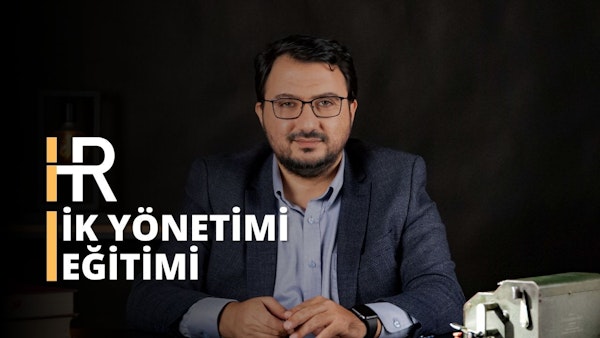 Bu görüntü, masa başında muhtemelen bir İnsan Kaynakları Yönetimi kursuna katılan bir adamı yakalıyor. Gözlüklü ve takım elbiseli adamın yakın çekimde gözleri, saati ve gömleği görülüyor. Gözlüklü ve sakallı yüzü görülüyor. Gömleğinin kolu da görülebiliyor. Masada metal bir kutu bulunmaktadır. Bu görüntü, İK sertifikasyon kurslarının önemini ve profesyonellerin bu alanda mükemmelleşmek için ihtiyaç duydukları beceri ve bilgileri kazanmalarına nasıl yardımcı olabileceklerini ideal bir şekilde temsil etmektedir. İK sertifikası, işverenlere çalışanlarının işi yapmak için gerekli niteliklere sahip olduğuna dair güven verir. Ayrıca iş arayanlara da seçtikleri meslekte mükemmelleşmek için doğru becerilere sahip olduklarına dair güvence sağlar.