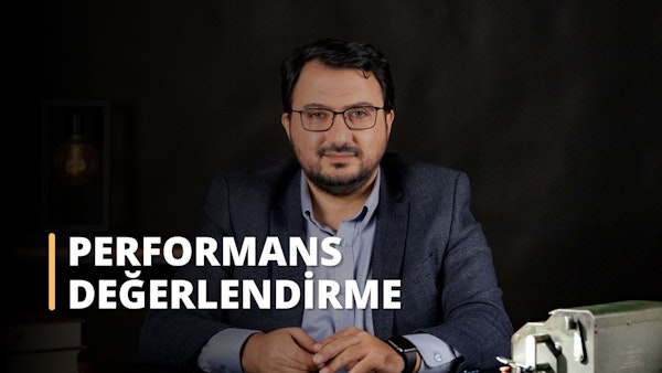 Bu görüntüde masa başında oturmuş performansını değerlendiren bir adam görülüyor. Takım elbise giyiyor ve masanın üzerinde metal bir kutu, bir saat ve yakın planda yüzü ve göğsü görünüyor. Adam açıkça kendini değerlendirdiği için bu görüntü performans değerlendirme eğitimiyle ilgili bir çevrimiçi kursa ait olabilir.