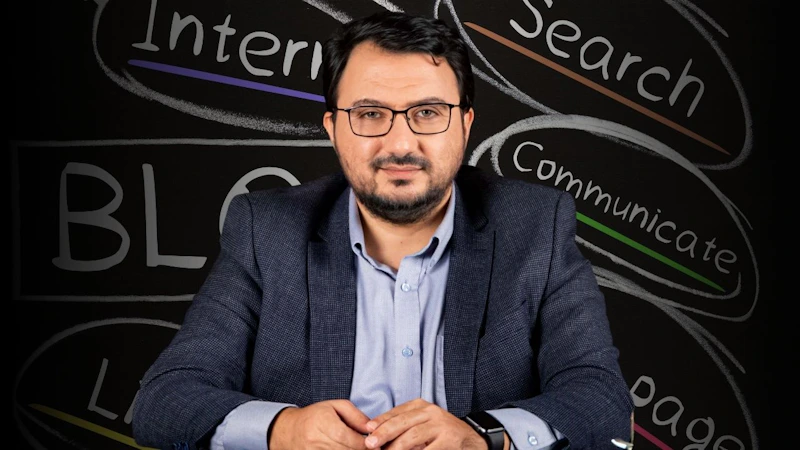 Bu, blog yazmayla ilgili bir çevrimiçi kursun görüntüsüdür. Takım elbiseli bir adam kara tahtanın önünde oturuyor, gözlüklü ve sakallı, bilgi ve otorite izlenimi veriyor. Konuşmacıyı dikkatle dinliyor gibi görünüyor. Tahtanın üzerinde siyah beyaz bir 'B' harfi var, muhtemelen blog yazmaya atıfta bulunuyor. Adamın üzerinde düğmesi yakından çekilmiş bir gömlek ve kolunda bir saat vardır. Gözleri de görülebiliyor, bu da dikkatle dinlediğini gösteriyor. Tüm bu ayrıntılar adamın bir blog kursuna katıldığına işaret ediyor. Bu görüntü sayesinde dijital çağda blog yazmanın önemini görebiliyoruz.