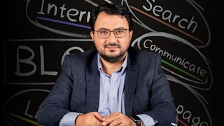 Bu, blog yazmayla ilgili bir çevrimiçi kursun görüntüsüdür. Takım elbiseli bir adam kara tahtanın önünde oturuyor, gözlüklü ve sakallı, bilgi ve otorite izlenimi veriyor. Konuşmacıyı dikkatle dinliyor gibi görünüyor. Tahtanın üzerinde siyah beyaz bir 'B' harfi var, muhtemelen blog yazmaya atıfta bulunuyor. Adamın üzerinde düğmesi yakından çekilmiş bir gömlek ve kolunda bir saat vardır. Gözleri de görülebiliyor, bu da dikkatle dinlediğini gösteriyor. Tüm bu ayrıntılar adamın bir blog kursuna katıldığına işaret ediyor. Bu görüntü sayesinde dijital çağda blog yazmanın önemini görebiliyoruz.