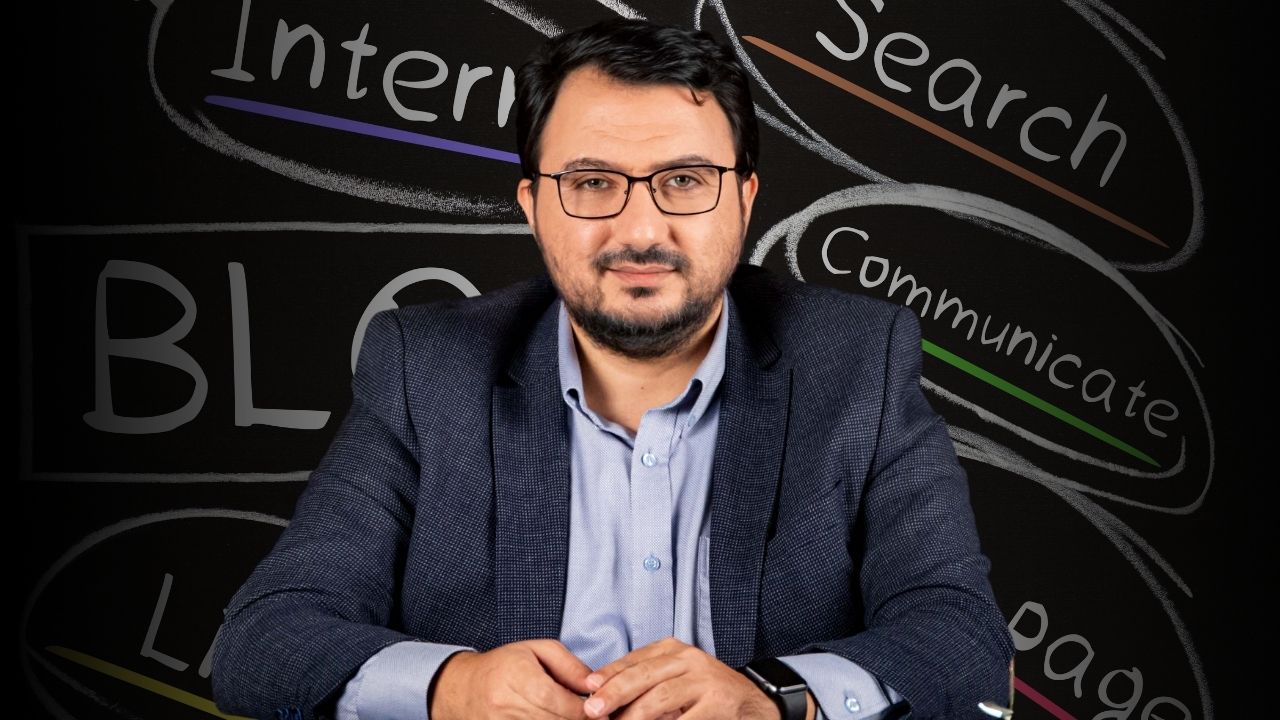 Bu, blog yazmayla ilgili bir çevrimiçi kursun görüntüsüdür. Takım elbiseli bir adam kara tahtanın önünde oturuyor, gözlüklü ve sakallı, bilgi ve otorite izlenimi veriyor. Konuşmacıyı dikkatle dinliyor gibi görünüyor. Tahtanın üzerinde siyah beyaz bir 'B' harfi var, muhtemelen blog yazmaya atıfta bulunuyor. Adamın üzerinde düğmesi yakından çekilmiş bir gömlek ve kolunda bir saat vardır. Gözleri de görülebiliyor, bu da dikkatle dinlediğini gösteriyor. Tüm bu ayrıntılar adamın bir blog kursuna katıldığına işaret ediyor. Bu görüntü sayesinde dijital çağda blog yazmanın önemini görebiliyoruz.