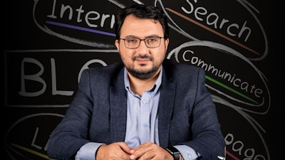 Bu, blog yazmayla ilgili bir çevrimiçi kursun görüntüsüdür. Takım elbiseli bir adam kara tahtanın önünde oturuyor, gözlüklü ve sakallı, bilgi ve otorite izlenimi veriyor. Konuşmacıyı dikkatle dinliyor gibi görünüyor. Tahtanın üzerinde siyah beyaz bir 'B' harfi var, muhtemelen blog yazmaya atıfta bulunuyor. Adamın üzerinde düğmesi yakından çekilmiş bir gömlek ve kolunda bir saat vardır. Gözleri de görülebiliyor, bu da dikkatle dinlediğini gösteriyor. Tüm bu ayrıntılar adamın bir blog kursuna katıldığına işaret ediyor. Bu görüntü sayesinde dijital çağda blog yazmanın önemini görebiliyoruz.