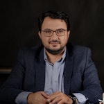 Bu, koyu mavi takım elbise giyen bir adamın fotoğrafı. Arkasında beyaz bir duvar olan parlak ışıklı bir odada durmaktadır. Adamın kısa siyah saçları var ve yüzünde ciddi bir ifadeyle önüne bakıyor. Beyaz bir elbise gömleği, mavi çizgili bir kravat ve koyu mavi bir takım elbise ceketi giymektedir. Ayrıca siyah elbise pantolonu ve siyah elbise ayakkabıları giymektedir. Elleri yanındadır ve sol bileğinde bir saat takılıdır. Adam kendinden emin ve sakin görünüyor.