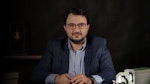 Bu, koyu mavi takım elbise giyen bir adamın fotoğrafı. Arkasında beyaz bir duvar olan parlak ışıklı bir odada durmaktadır. Adamın kısa siyah saçları var ve yüzünde ciddi bir ifadeyle önüne bakıyor. Beyaz bir elbise gömleği, mavi çizgili bir kravat ve koyu mavi bir takım elbise ceketi giymektedir. Ayrıca siyah elbise pantolonu ve siyah elbise ayakkabıları giymektedir. Elleri yanındadır ve sol bileğinde bir saat takılıdır. Adam kendinden emin ve sakin görünüyor.