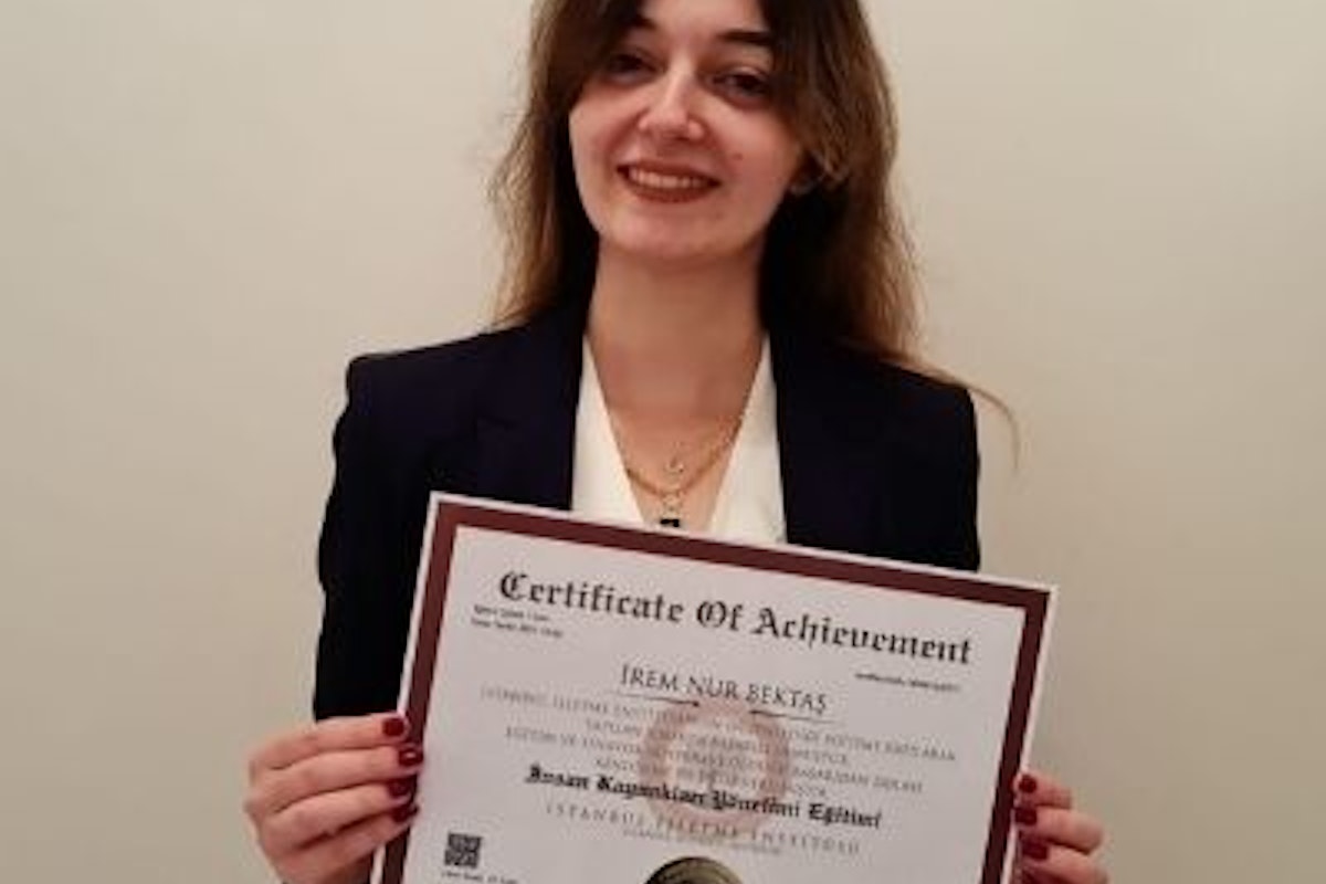 This photo displays a smiling woman holding a certificate in her hands. She has long dark hair that is pulled back into a neat bun, and wears a white blouse with a black skirt. The certificate is white and green with a gold seal and text on the front. She has a look of pride and accomplishment on her face, likely due to receiving the certificate. The background is a neutral off-white wall, with a few other certificates hung up behind her. This image could symbolize the feelings of success and achievement that come with a hard-earned accomplishment.