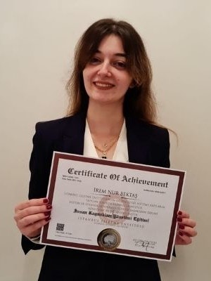 This photo displays a smiling woman holding a certificate in her hands. She has long dark hair that is pulled back into a neat bun, and wears a white blouse with a black skirt. The certificate is white and green with a gold seal and text on the front. She has a look of pride and accomplishment on her face, likely due to receiving the certificate. The background is a neutral off-white wall, with a few other certificates hung up behind her. This image could symbolize the feelings of success and achievement that come with a hard-earned accomplishment.