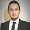 Şık gri takım elbiseli ve mavi kravatlı bir adam düz beyaz bir duvarın önünde duruyor. Elleri ceplerinde ve yüzünde ciddi bir ifade var. Saçları siyah ve düzgünce geriye taranmış. Takım elbisesinin altına beyaz bir gömlek giymiş ve beline siyah bir kemer bağlamış. Fotoğrafın ışığı arkasındaki duvarda hafif bir gölge yaratarak görüntüye derinlik katıyor. Adamın duruşu otorite ve güven hissi veriyor.