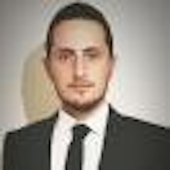 Şık gri takım elbiseli ve mavi kravatlı bir adam düz beyaz bir duvarın önünde duruyor. Elleri ceplerinde ve yüzünde ciddi bir ifade var. Saçları siyah ve düzgünce geriye taranmış. Takım elbisesinin altına beyaz bir gömlek giymiş ve beline siyah bir kemer bağlamış. Fotoğrafın ışığı arkasındaki duvarda hafif bir gölge yaratarak görüntüye derinlik katıyor. Adamın duruşu otorite ve güven hissi veriyor.