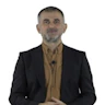 Takım elbiseli adam düz, beyaz bir arka planın önünde durmaktadır. Siyah, iki parçalı bir takım elbise ve açık mavi, uzun kollu bir gömlek giymektedir. Elleri önünde kenetlenmiş ve kendinden emin bir ifadeyle dümdüz karşıya bakıyor. Siyah saçları kısa kesilmiş ve düzgünce taranmış. Rahat ama profesyonel bir duruş sergiliyor. Sol kolunda bir saat ve siyah deri bir kemer takıyor. Görünüşü sofistike ve dengeli.