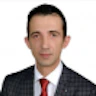 Bu, şık siyah bir takım elbise giyen ve lacivert kravat takan orta yaşlı yakışıklı bir adamın fotoğrafı. Adamın yüzünde hoş bir ifade var ve saçları düzgünce taranmış. Takım elbisesi iyi dikilmiş ve üzerine tam oturmuş. Takım elbisenin altına bembeyaz bir gömlek giymiş ve kravatı boynunda sıkıca düğümlenmiş. Ellerini önünde rahatça kavuşturmuş, kendinden emin ve kendinden emin bir duruş sergiliyor. İşlerini verimli ve etkili bir şekilde halletmeye alışkın birine benziyor. Profesyonellik ve başarı timsalidir.
