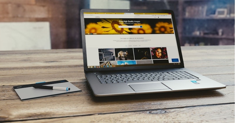 Beyaz bir masa üzerine yerleştirilmiş bir dizüstü bilgisayar görüntünün odak noktasıdır. Arka planda bir kişinin yüzü hafifçe görülebiliyor, görüntüde bulanık. Dizüstü bilgisayar ekranında görüntüler ve metinlerle birlikte bir bilgisayarın ekran görüntüsü var. Ön planda, gümüş bir kalem bir tabletin üstünde duruyor ve siyah tuşlu beyaz dikdörtgen bir nesne yakınlarda. Tuşları standart bir düzende dizilmiş bir klavyenin yakın çekimi de görülüyor. Son olarak, görüntünün sağ üst kısmında koyu renk saçlı ve kırmızı rujlu bir kadın görülüyor.