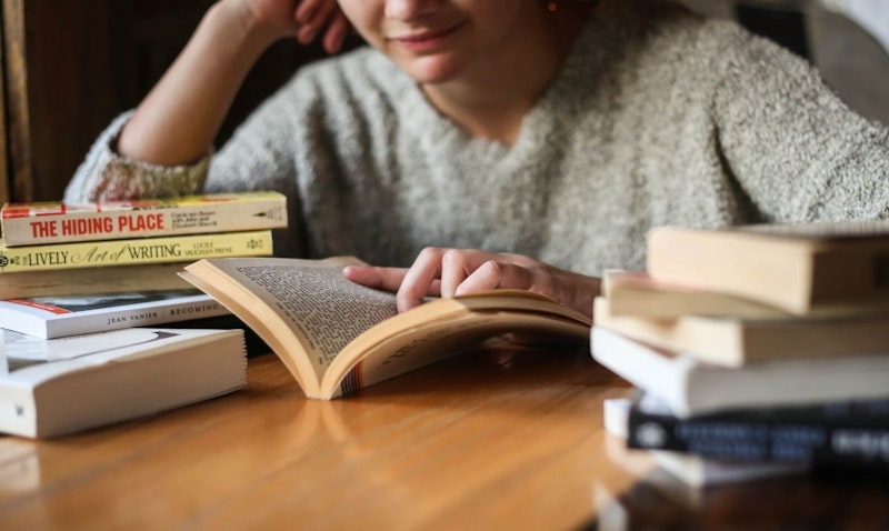 Bir kişi elinde açık bir kitapla sandalyede oturmaktadır. Kitap kişiye yakın tutularak kapağının ve bazı sayfalarının net bir şekilde görülmesi sağlanmıştır. Kişinin bir eli kitabın kapağında, diğer eli ise sayfaları çevirmektedir. Kişi kitaba dalmış, gözleri önündeki sayfaya odaklanmış gibi görünüyor. Arka plan bulanıktır ve görüntünün rüya gibi bir niteliği vardır. Görüntü yakın çekim olduğundan kitabın tüm detayları görülebiliyor ve kişinin yüz hatları hafifçe bulanık. Kişi kitaptan keyif alıyor gibi görünmektedir ve görüntünün genel atmosferi huzurlu ve sakinleştiricidir.