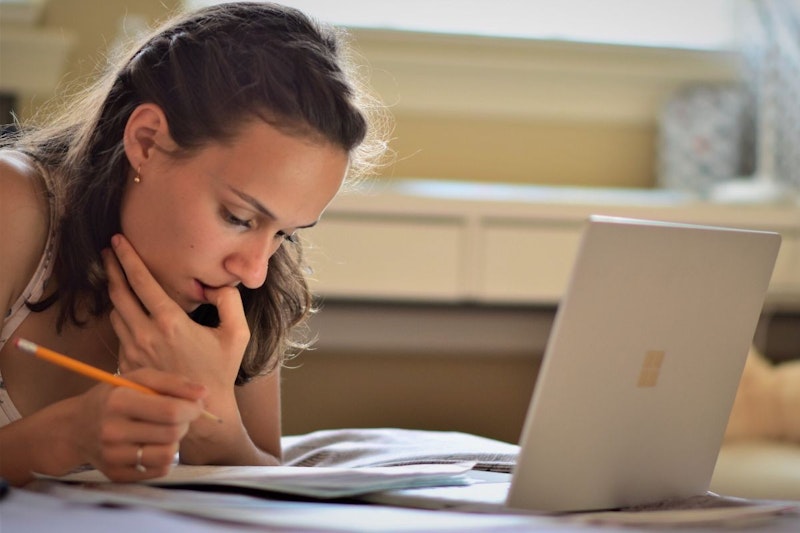 Genç bir kadın bir dizüstü bilgisayarın önünde oturuyor, yüzü parlak ekran tarafından aydınlatılıyor. Üzerinde gri bir tişört ve mavi bir kot pantolon var ve uzun kahverengi saçları arkaya doğru toplanmış. Sağ elinde bir kalem tutuyor ve dikkatle yere bakıyor, gözleri ekrana odaklanmış. Sol eli sanki bir sonraki adımda ne yapacağına karar veriyormuş gibi mousepad'in üzerinde duruyor. Dizüstü bilgisayar, çarşafları hafifçe buruşmuş bir yatağın üzerinde duruyor ve odayı sıcak, sade bir atmosfer dolduruyor. Kadın kendini tamamen işine kaptırmış, konsantrasyonu hiç bozulmamış.