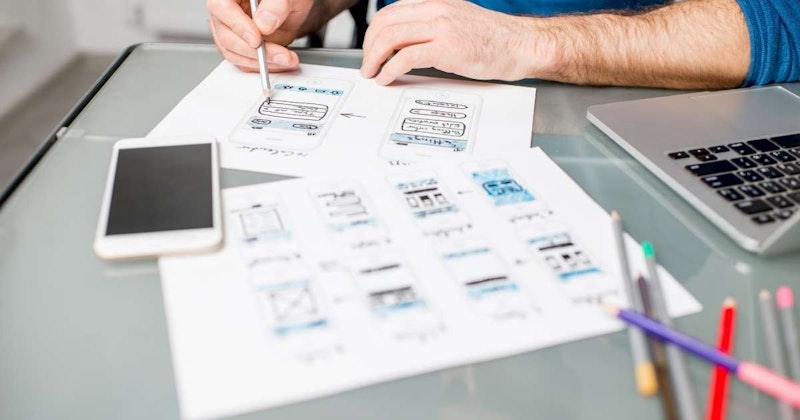 Bir kâğıt parçası üzerine eskiz çizen bir kişinin yakın çekimi. Kişi eskiz çizmek için görüntüde görünen sağ elini kullanmaktadır. Elinde bir kalem tutmaktadır ve kalem kağıda eskiz çizmek için kullanılmaktadır. Kağıt beyaz ve kalem grinin açık bir tonunda. Kişinin yüzü kısmen görülebiliyor ve çizimine konsantre olmuş gibi görünüyor. Resmin arka planında birkaç kalem, bir telefon ve bir dizüstü bilgisayar klavyesi var. Telefon siyah, dizüstü bilgisayar klavyesi ise gümüş rengindedir. Kişi odaklanmış görünüyor ve çizimine çok dikkat ediyor.