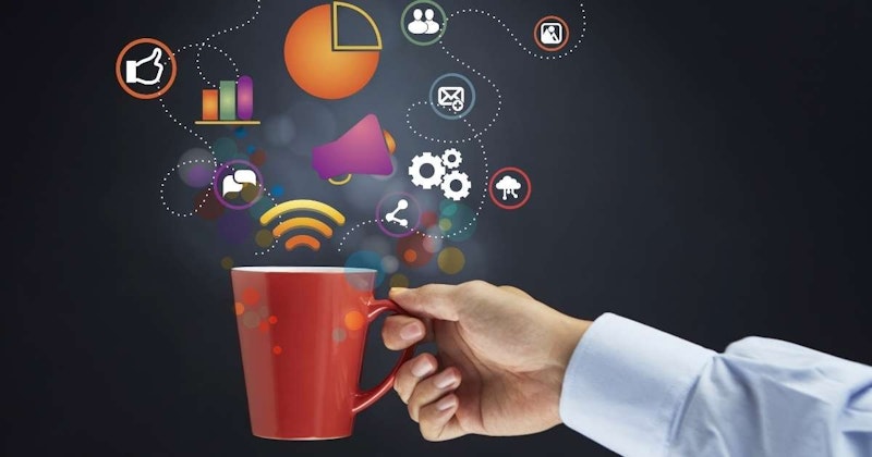 Üzerinde karmaşık simgeler bulunan kırmızı bir kupayı tutan bir elin yakın çekimi. Kupanın beyaz bir kulpu var. Kupanın üzerindeki simgeler renkli ve dikdörtgen şeklinde, daire şeklinde dizilmiş. Dairenin ortasında beyaz bir başparmak yukarı işareti var. Yakınında içinde yarım daire olan turuncu bir daire vardır. Resmin arka planı koyu renklidir.