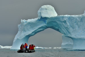 Bir teknedeki bir grup insan devasa bir buzdağının altındadır. Tekne beyaz ve mavi bir buz kemeriyle çevrilidir ve insanların parlak kırmızı ve mavi ceketleri kara karşı göze çarpmaktadır. Kırmızı montlu bir kişi kendi fotoğrafını çekerken, bir diğeri de ön planda kırmızı mont giymektedir. Arka planda bir motorun yakın çekimi ve uzaktaki bir buzdağının yakın çekimi görülüyor. Sahne, görkemli buzdağının altındaki tekne ve insanlarla güzel ve dingin.