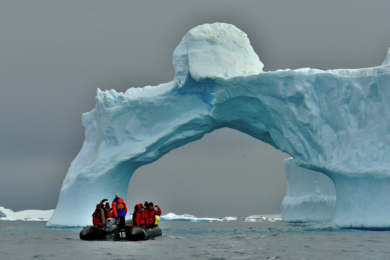 Bir teknedeki bir grup insan devasa bir buzdağının altındadır. Tekne beyaz ve mavi bir buz kemeriyle çevrilidir ve insanların parlak kırmızı ve mavi ceketleri kara karşı göze çarpmaktadır. Kırmızı montlu bir kişi kendi fotoğrafını çekerken, bir diğeri de ön planda kırmızı mont giymektedir. Arka planda bir motorun yakın çekimi ve uzaktaki bir buzdağının yakın çekimi görülüyor. Sahne, görkemli buzdağının altındaki tekne ve insanlarla güzel ve dingin.