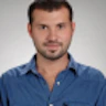 Parlak mavi gömlekli bir adam düz bir arka planın önünde duruyor. Kısa siyah saçları ve temiz traşlı bir yüzü vardır. Elleri arkasında, kameradan uzağa bakmaktadır. Vücudu hafifçe yana dönüktür ve izleyiciye neredeyse profilden bir görünüm verir. Üzerinde beyaz cepli açık mavi yakalı bir gömlek ve koyu mavi bir kot pantolon var. Duruşu rahat ve gevşek bir tavır sergilediğini gösteriyor. İfadesi memnuniyet ve huzur dolu. Gömleğinin mavisi nötr arka planla güzel bir tezat oluşturarak görsel açıdan hoş bir görüntü yaratıyor.