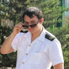 Kadrajdaki adam altın düğmeli lacivert bir blazer ceket, beyaz bir elbise gömleği ve siyah bir kravattan oluşan şık bir üniforma giyiyor. Saçları kısa kesilmiş ve düzgünce taranmış. Göğsü kabarık, omuzları geride ve kolları sert bir şekilde iki yanında duruyor. Yüz ifadesi ciddi ve bakışları uzaktaki bir şeye odaklanmış. Gururlu ve vakur görünüyor, askeri görev ve onurun bir sembolü. Sadakat, sorumluluk ve adanmışlığın önemini hatırlatıyor.