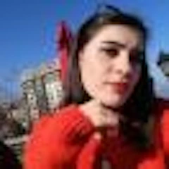 Genç bir kadın düz bir arka planın önünde duruyor. Üzerinde bileklerine kadar inen uzun kollu, parlak kırmızı bir gömlek var. Saçları kahverenginin koyu bir tonunda ve yüzünde ciddi bir ifade var. Gözleri kameradan uzağa bakıyor ve dudakları hafifçe ayrılmış. Kollarını vücuduna yakın tutuyor ve duruşu kendinden emin ama rahat. İnce bir yapısı var ve duruşu düzgün. Görüntünün odak noktası o ve varlığı güçlü ve kuvvetli.