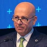 Takım elbiseli adam yüzünde ciddi bir ifadeyle dimdik ayakta duruyor. Açık mavi takım elbisesi tam oturmuş ve aynı derecede net beyaz bir gömlek ve koyu mavi kravatla tamamlanmış. Saçları düzgünce geriye taranmış ve gözleri uzaktaki bir şeye odaklanmış. Parlatılmış siyah deri elbise ayakkabıları giyiyor. Elleri önünde birbirine kenetlenmiş ve bir şey bekliyor gibi görünüyor. Duruşu kendinden emin ve ağırbaşlı, profesyonellik ve yetkinlik havası taşıyor. Önüne çıkan her türlü zorluğun üstesinden gelmeye hazır görünüyor.