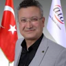 İyi giyimli, gözlüklü bir adam aydınlık bir odada durmaktadır. Üzerinde siyah bir takım elbise, altında beyaz bir gömlek ve kalın çerçeveli bir gözlük var. Saçları düzgünce şekillendirilmiş ve nötr bir ifadeyle dümdüz karşıya bakıyor. Elleri hafifçe arkada, ayakta duruyor ve duruşu kendinden emin ve sakin. Otuzlu yaşlarının ortalarında ve orta yapılı görünüyor. Bakışları odaklanmış ve yoğun, yüz hatları keskin ve iyi tanımlanmış. Kendisi saygın bir profesyonel ve bu görüntü bir güven ve ciddiyet hissi veriyor.