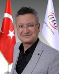 İyi giyimli, gözlüklü bir adam aydınlık bir odada durmaktadır. Üzerinde siyah bir takım elbise, altında beyaz bir gömlek ve kalın çerçeveli bir gözlük var. Saçları düzgünce şekillendirilmiş ve nötr bir ifadeyle dümdüz karşıya bakıyor. Elleri hafifçe arkada, ayakta duruyor ve duruşu kendinden emin ve sakin. Otuzlu yaşlarının ortalarında ve orta yapılı görünüyor. Bakışları odaklanmış ve yoğun, yüz hatları keskin ve iyi tanımlanmış. Kendisi saygın bir profesyonel ve bu görüntü bir güven ve ciddiyet hissi veriyor.