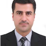 Şık giyimli bir adam, üç parçalı takım elbisesi ve kravatıyla kendinden emin bir şekilde duruyor. Siyah bir takım elbise, beyaz bir gömlek ve kırmızı bir kravat giymiştir. Adamın düzgünce taranmış siyah saçları ve temiz traşlı bir yüzü vardır. Elleri önünde kenetlenmiş ve yüzünde hafif bir gülümsemeyle ileriye bakıyor. Solunda bir pencere bulunan gri bir duvarın önünde duruyor ve dışarıdan gelen ışık siluetine gölge düşürüyor. Duruşu güç, güven ve gurur duygusu yayıyor.