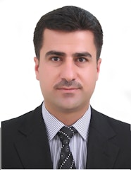 Şık giyimli bir adam, üç parçalı takım elbisesi ve kravatıyla kendinden emin bir şekilde duruyor. Siyah bir takım elbise, beyaz bir gömlek ve kırmızı bir kravat giymiştir. Adamın düzgünce taranmış siyah saçları ve temiz traşlı bir yüzü vardır. Elleri önünde kenetlenmiş ve yüzünde hafif bir gülümsemeyle ileriye bakıyor. Solunda bir pencere bulunan gri bir duvarın önünde duruyor ve dışarıdan gelen ışık siluetine gölge düşürüyor. Duruşu güç, güven ve gurur duygusu yayıyor.